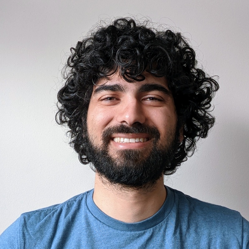 Abdul smiling, with curly black hair and a beard, in a blue T-shirt against a plain wall
